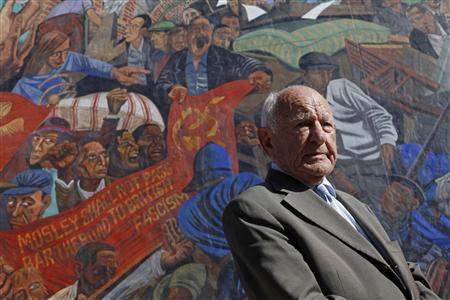 Max Levitas poses for a photograph in front of a mural depicting the 1936 battle of Cable Street, on Cable Street, in east London